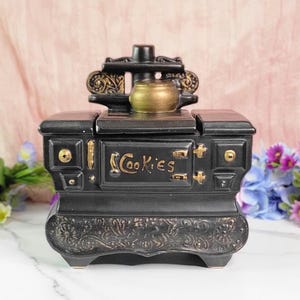 May include: Black ceramic cookie jar shaped like a vintage wood-burning stove with gold accents and the word "Cookies" written on the front.