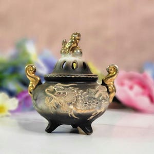 May include: A black and gold ceramic incense burner with a dragon design and a small golden figure on top. The burner has three legs and two handles.