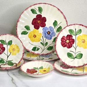 May include: A collection of cream-colored ceramic plates and bowls with a floral design. Each piece features a hand-painted flower in shades of red, yellow, and blue, with green leaves and a scalloped, red-trimmed edge. The set includes various sizes.