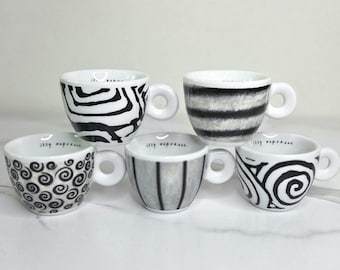 May include: Five white espresso cups with black and white patterns. Designs include zebra stripes, horizontal bands, vertical stripes, and swirling patterns. Each cup has a round handle and the words "illy espresso" inside.