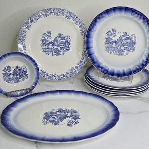 May include: A collection of vintage blue and white porcelain dishes. The set includes plates, bowls, and an oval platter. Each piece features a detailed blue landscape design on a white background, with decorative blue rims.