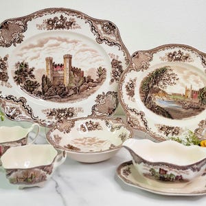 May include: A set of brown and white ceramic dishes with a floral pattern and a castle scene. The set includes a large oval platter, a smaller oval platter, a round bowl, and two gravy boats.