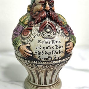 May include: A ceramic beer stein shaped like a bearded man wearing a green hat and apron. The apron has text in German. The stein is primarily beige, with accents of green, purple, and red.