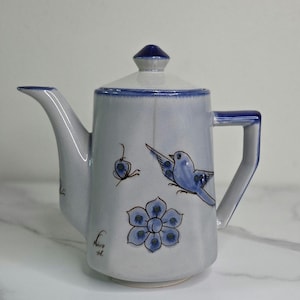 May include: A ceramic teapot with a lid, spout, and handle. The teapot is light gray with blue accents and hand-painted blue bird and floral designs. The teapot has a signature on the side.