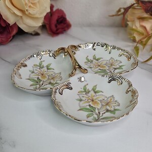 May include: A three-sectioned, white porcelain serving dish with a gold handle and trim. Each section features a floral design with yellow and green accents. The dish is ideal for serving appetizers or small treats.
