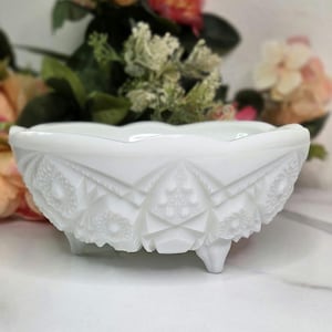 May include: A white, footed, decorative bowl with a scalloped rim. The bowl features a detailed, embossed pattern of geometric shapes and floral designs. The bowl is made of opaque white glass and is likely vintage.