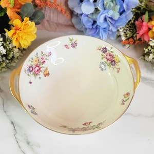 May include: A white porcelain serving bowl with gold trim and a floral pattern. The bowl has two gold handles.