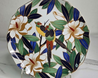 May include: Decorative ceramic plate with a scalloped edge, featuring a design of two colorful birds perched on a branch amidst white flowers and green and blue leaves. The plate has a gold rim and is displayed on a clear stand.
