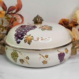 May include: A white enamel casserole dish with a lid, featuring grape cluster and leaf designs in purple, green, and gold. The dish has gold-colored handles and a decorative knob on the lid. The rim is silver-colored.