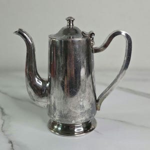 May include: A silver-colored coffee pot with a curved spout and handle. The pot has a lid with a small knob on top and sits on a rounded base. The surface shows signs of wear.