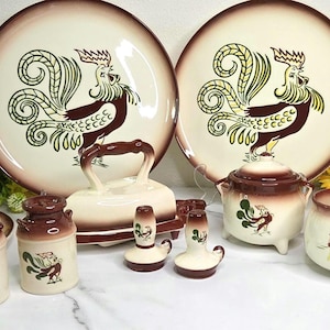 May include: A ceramic dinnerware set featuring a cream base with brown rim accents and rooster illustrations. The collection includes two large plates, a butter dish with a handle, a lidded sugar bowl, a creamer, two shakers, and two small canisters.
