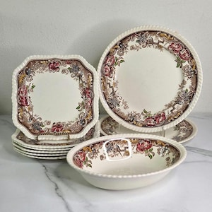 May include: A collection of vintage dinnerware featuring a cream-colored base with a floral and vine pattern in shades of brown, red, and green. The set includes plates and a bowl with a scalloped edge. The plates are stacked on a gold-colored plate stand.