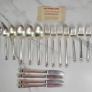 May include: A collection of vintage silver-plated flatware, including spoons, forks, and knives, arranged on a marble surface. The silverware features ornate floral detailing on the handles. A guarantee card is visible in the background.
