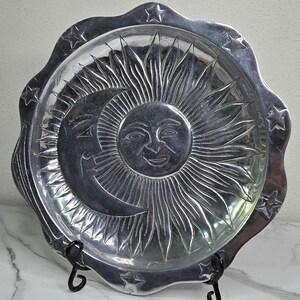 May include: A silver-colored decorative plate with a celestial design. The plate features a smiling sun and crescent moon in the center, surrounded by radiating lines and star-shaped accents along the scalloped edge. The plate is displayed on a black metal stand.