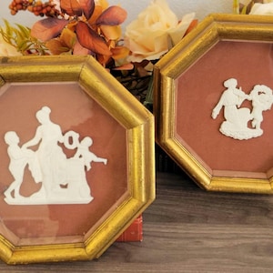 May include: Two octagonal frames with gold trim and a red background. Each frame contains a white ceramic cameo depicting a scene with figures.