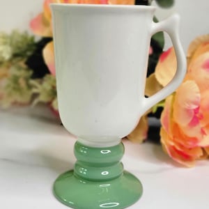 May include: A white ceramic mug with a green pedestal base. The mug has a curved handle and a smooth, glossy finish. The base has a rounded shape with a series of decorative rings. The mug is likely for beverages.