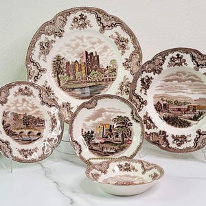 May include: A set of brown and white ceramic dinnerware with a castle and floral pattern. The set includes a large dinner plate, a smaller dinner plate, a soup bowl, and a small bowl.