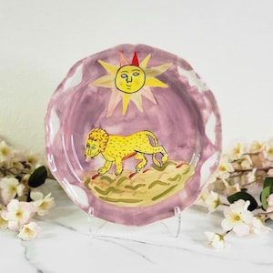 May include: A ceramic plate with a pink background and a yellow lion walking towards a yellow sun with a face. The sun has a red crown and the plate has a scalloped edge.
