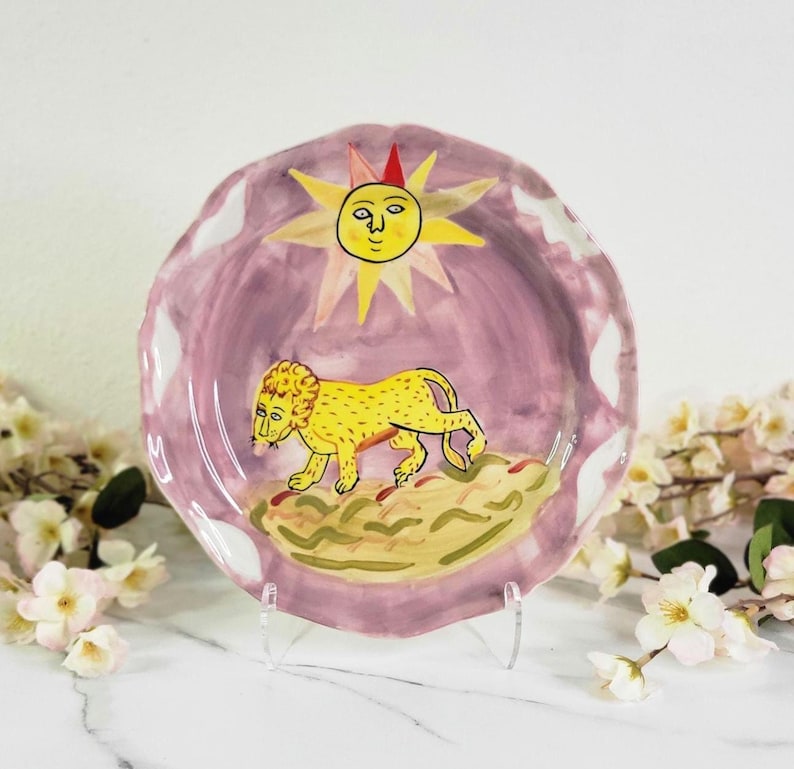 May include: A ceramic plate with a pink background and a yellow lion walking towards a yellow sun with a face. The sun has a red crown and the plate has a scalloped edge.