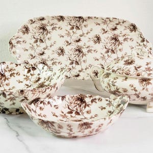 May include: A set of four white ceramic serving dishes with a brown floral pattern. The set includes a large oval platter, a round bowl, a smaller oval bowl, and a covered serving dish.