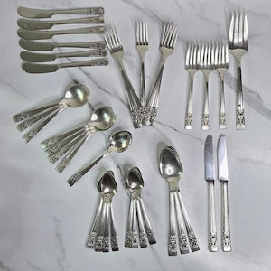 May include: A collection of vintage silver-plated flatware, including butter knives, forks, spoons, and teaspoons. The silverware features ornate floral detailing on the handles. The arrangement is displayed on a marble-patterned surface.