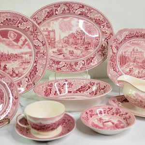 May include: A collection of antique pink and white china dishes, including plates, bowls, a cup and saucer, and a gravy boat. Each piece features a detailed landscape scene and decorative border.