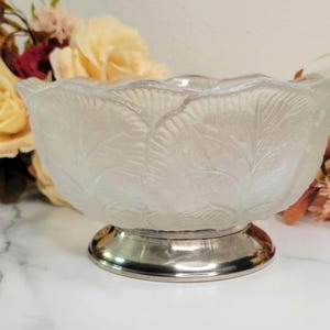 May include: A frosted glass bowl with a leaf pattern, set on a silver-colored base. The bowl has a scalloped rim and is surrounded by flowers in the background. The bowl is likely used for serving or display.
