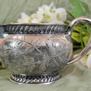 May include: A silver creamer with a floral design. The creamer has a round body and a handle. The floral design is embossed on the surface of the creamer.