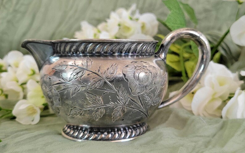 May include: A silver creamer with a floral design. The creamer has a round body and a handle. The floral design is embossed on the surface of the creamer.