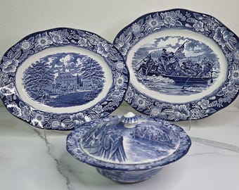 May include: A set of blue and white ceramic serving dishes. The set includes two oval platters and a covered bowl. The platters and bowl feature floral borders and historical scenes.