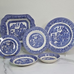 May include: A collection of blue and white Willow pattern china dishes. The set includes various plates, a serving dish, and a bowl. The plates have a white center with a blue landscape design and a blue border. The serving dish has a scalloped edge.