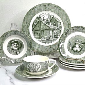 May include: A vintage green and white dinnerware set featuring plates, bowls, a teacup, and saucers. The plates have a decorative border and center illustrations of a building, a man with a pipe, and a teapot. The set includes multiple pieces.