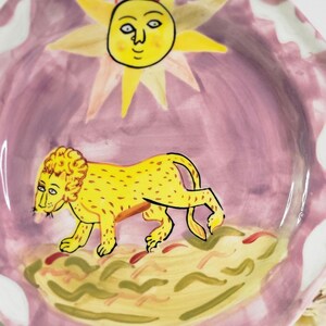 May include: A pink ceramic plate with a yellow sun and a yellow lion walking on a green and yellow ground. The sun has a face and the lion has a mane.