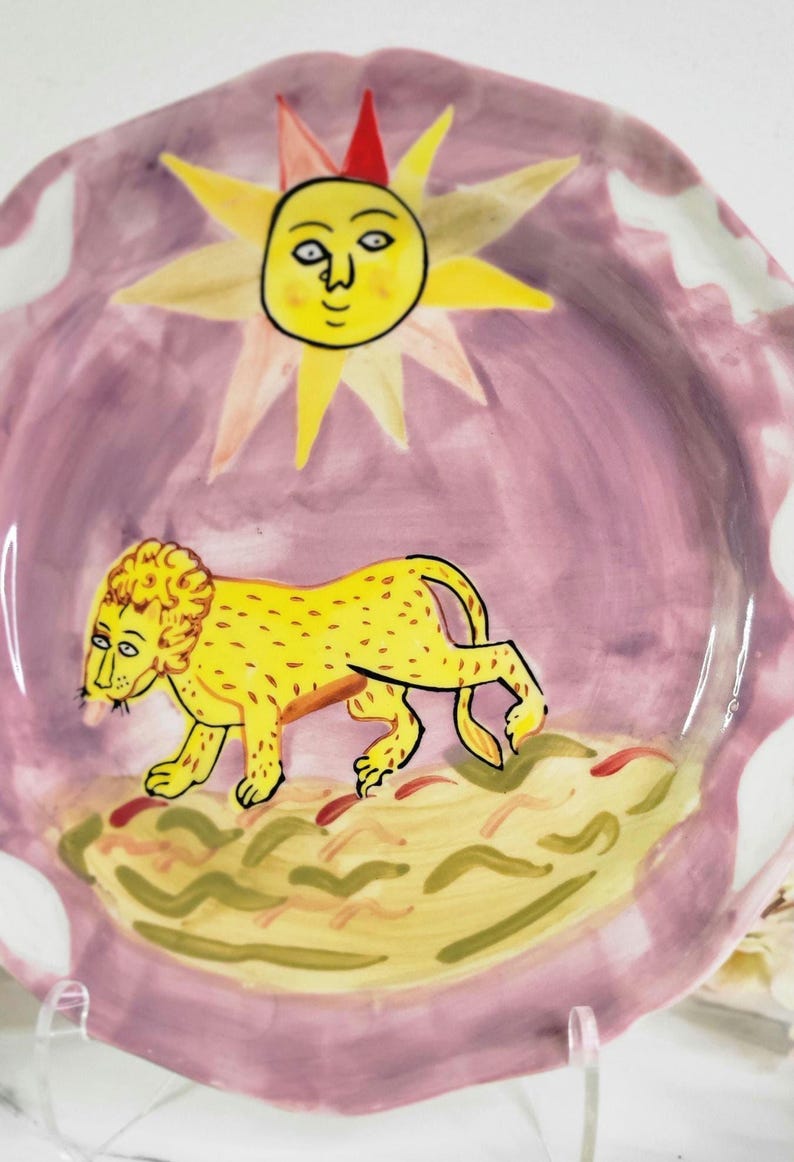 May include: A pink ceramic plate with a yellow sun and a yellow lion walking on a green and yellow ground. The sun has a face and the lion has a mane.