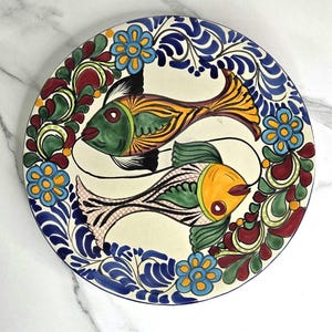May include: A round ceramic plate featuring a cream-colored background with two stylized fish swimming in a circle. The fish are painted in shades of green, yellow, and orange. The border is decorated with blue flowers and swirling green and red foliage.
