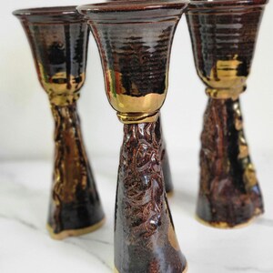 May include: Set of three brown ceramic cups with gold accents. The cups have a textured surface and a flared rim. The cups are arranged in a row on a white surface.