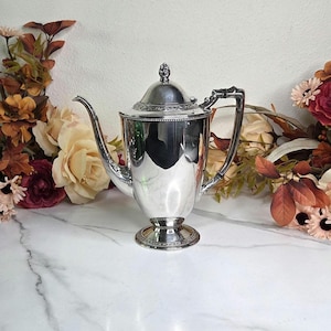 May include: A silver-colored coffee pot with a lid and a handle. The pot has a long spout and sits on a pedestal base. The background features floral arrangements with roses and other flowers.