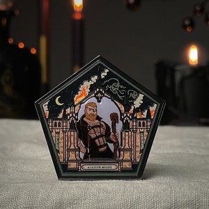 May include: A black and gold enamel pin shaped like a Hogwarts house crest. The crest features a wizard with a beard and a staff, a burning building, and the text "ALASTOR MOODY".