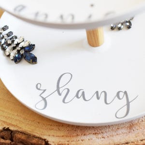 May include: White two-tiered serving tray with a wooden support. The word "zhang" is written in cursive on the bottom tier. The top tier has the word "annie" written on it. Embellishments include blue and clear crystal jewelry.