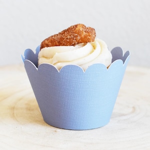 May include: A cupcake with white frosting and a cinnamon sugar pastry topping, presented in a light blue scalloped cupcake liner. The cupcake sits on a light wooden surface.