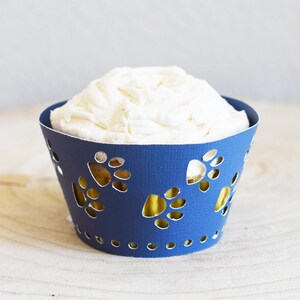 Puede incluir: Un envoltorio de cupcake azul con recortes en forma de huella de pata. El envoltorio está decorado con glaseado blanco.