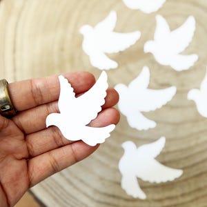 May include: White dove-shaped cutouts with outstretched wings are scattered on a wooden surface. One cutout is held in a hand, with others visible in the background. The doves are a bright white color.
