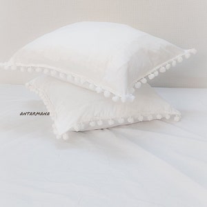 May include: Two white pillowcases with pom-pom trim. The pillowcases are stacked on a white bedspread. The text "ANTARMANA" is visible on the bottom pillowcase.