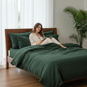 Puede incluir: Una cama con una funda nórdica verde oscuro, fundas de almohada y una sábana bajera. La cama tiene un marco de madera. Una persona está sentada en la cama leyendo un libro. La habitación tiene una ventana grande con cortinas blancas y una planta en maceta.