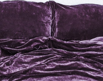 Puede incluir: Un edredón de terciopelo morado con dos fundas de almohada a juego. El edredón está arrugado y tiene un aspecto suave y lujoso.
