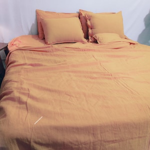 May include: A yellow linen bedding set with a flat sheet, duvet cover, and two pillowcases. The bedding is arranged on a bed with a wooden headboard.