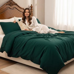 May include: A bed with a dark green duvet cover and matching pillowcases. A decorative pillow with a beige woven design rests on the bed. The headboard is made of dark wood. A person wearing white pajamas is lying on the bed.