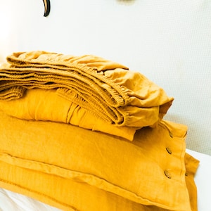 May include: A stack of mustard yellow linen bedding with two pillowcases and a fitted sheet. The fitted sheet has elastic around the edges. The pillowcases have buttons on the edges. The bedding is folded neatly on a white surface.