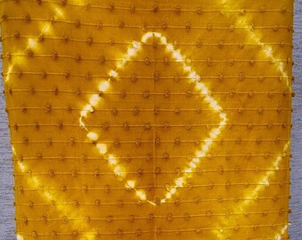Puede incluir: Una manta de color amarillo mostaza con un patrón de tie-dye blanco y flecos. La manta tiene una superficie texturizada con pequeños bultos en relieve.