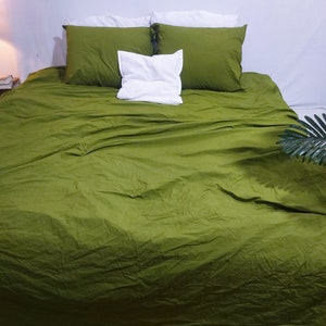 May include: A bed dressed in olive green bedding, including a fitted sheet, two pillowcases, and a white pillow. The bed has a white bed skirt. The wall behind the bed is white.